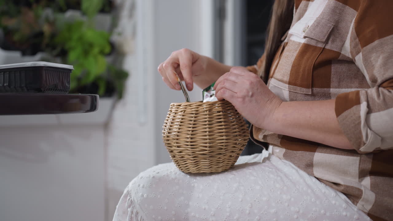Lower view of horticulturist seated on sofa near round table placing paper seed cards into small woven basket on lap amidst indoor potted plants and cozy white brick backdrop under soft natural light