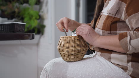 lower view of horticulturist seated on sofa near round table placing paper seed cards into small woven basket on lap amidst indoor potted plants and cozy white brick backdrop under soft natural light