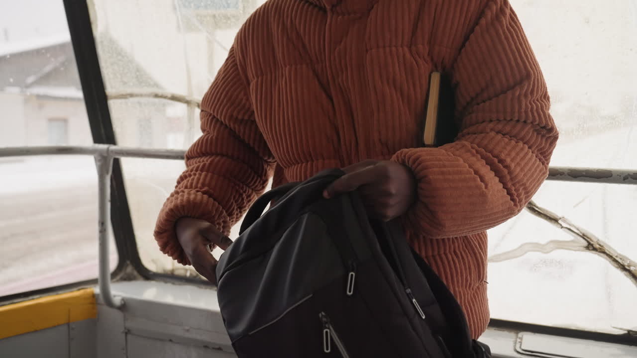 Black man, orange jacket, securing backpack inside vintage city bus cautious commuter checks zippers, protective posture, closeup hands, textured knit, cold breath on window, muted winter palette
