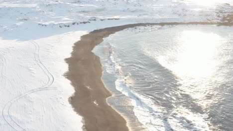 Bright-sunlight-shines-onto-winter-coastline-of-Iceland-with-waves,-aerial
