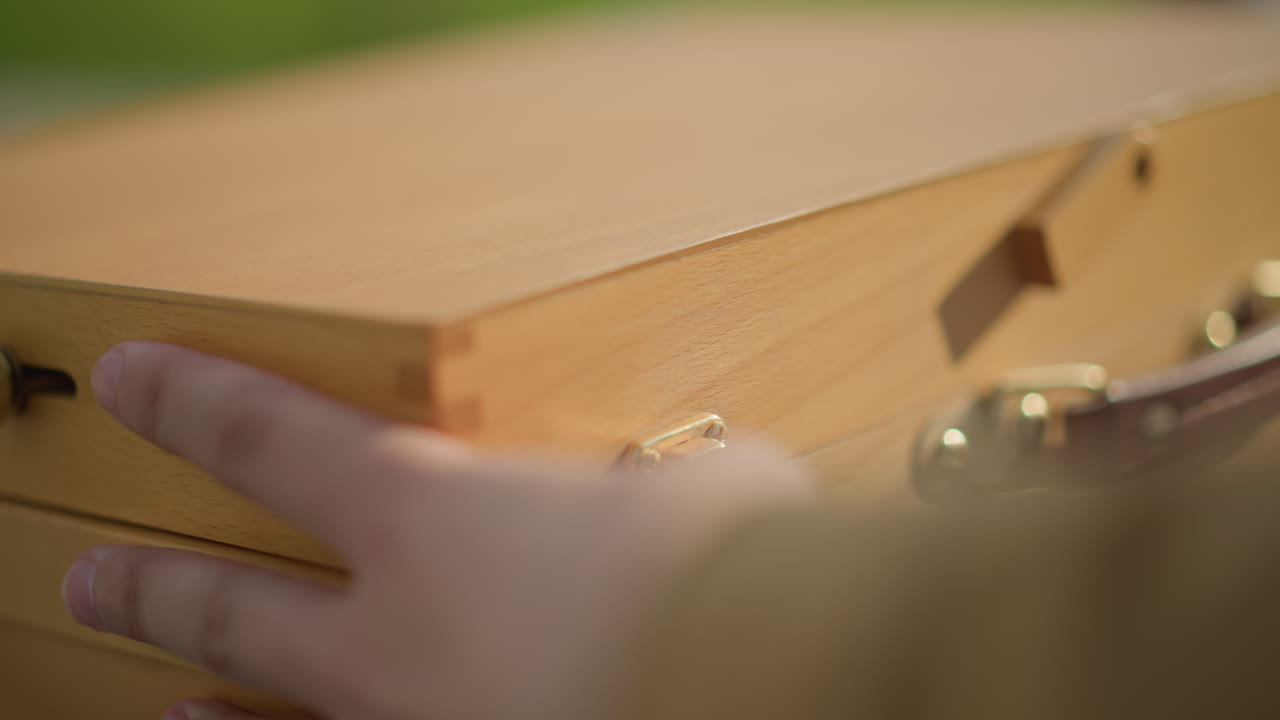 custodia di legno con colori su una panchina, primo piano di mani che sbloccano un fermo in ottone e un manico in pelle, luce solare che illumina le venature del legno, artista che prepara gli strumenti per uno schizzo per dipingere sulla strada, atmosfera tranquilla e concentrata