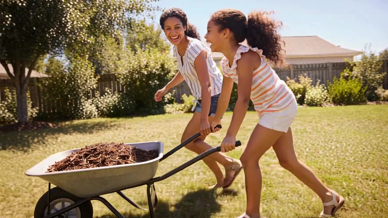 Grabbing handles starting mom and daughter in shorts pushing wheelbarrow in yard moving mulch