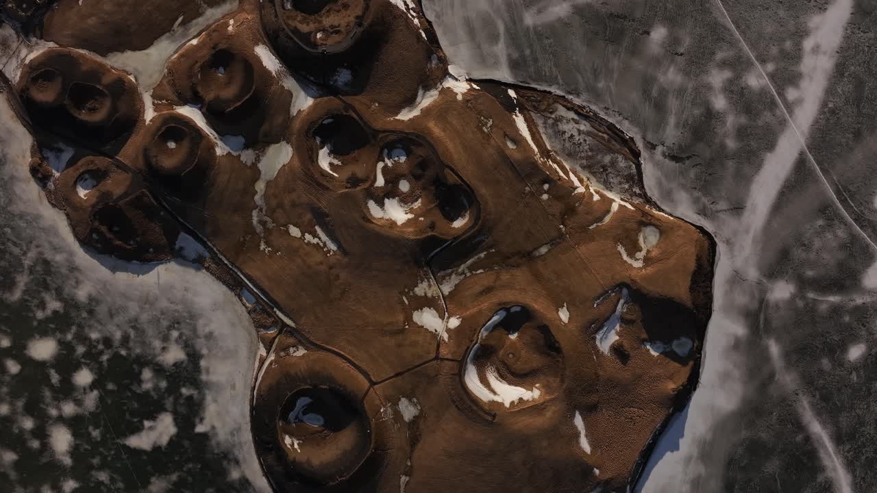 Top-down aerial view of frozen Lake Mývatn revealing volcanic pseudocraters near Skútustaðir, Iceland. Stark patterns and snow traces create an otherworldly geological texture.