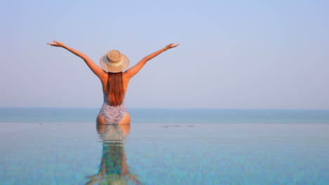 Back-of-exotic-woman-sitting-on-infinity-pool-border-and-raising-hands,-tropical-happiness-on-vacation,-slow-motion-full-frame