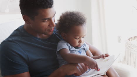 side view of millennial african american father sitting and reading a book with his toddler son, close up