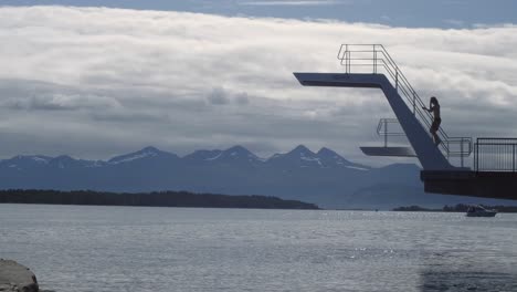 wide scenic shot of long haired, blonde, caucasian male climbing and jumping from diving tower doing a backflip gainer into water in profile in norway