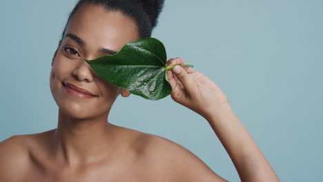 Porträt-Einer-Schönen-Afroamerikanischen-Frau,-Die-Ihr-Gesicht-Mit-Einem-Blatt-Berührt,-Einen-Glatten,-Gesunden-Teint-Streichelt-Und-Verspielte-Natürliche-Schönheit-Auf-Blauem-Hintergrund-Genießt.-Hautpflegekonzept