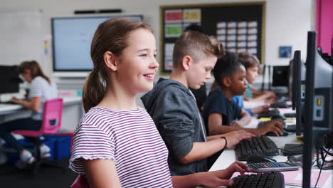 Female-Pupil-In-Line-Of-High-School-Students-Working-at-Screens-In-Computer-Class-With-Teacher-In-Background