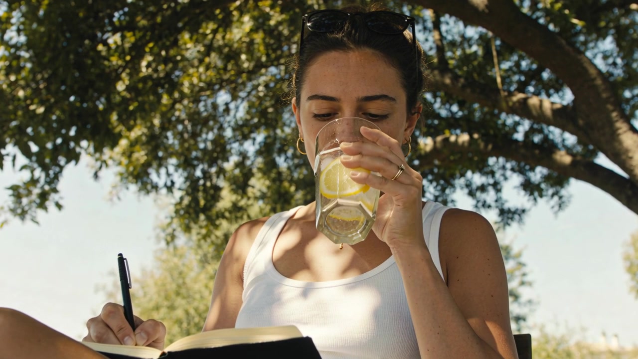 Mulher de regata pausando a escrita, levantando copo e bebendo para se refrescar, retomando caderno com caneta debaixo da árvore.