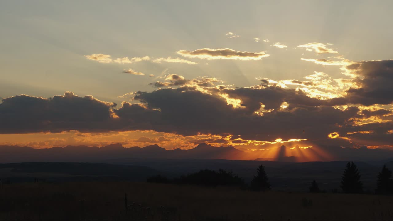 Beautiful sunbeams peak through clouds rolling past a sunset at the edge of the Alberta foothill; static shot 1080p