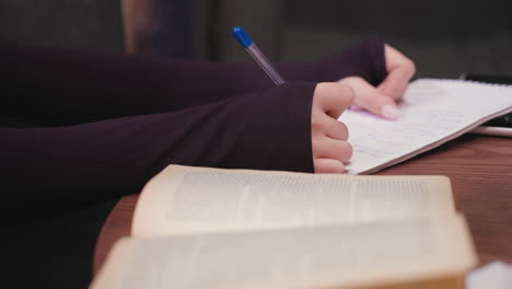 close up of woman hand holding blue pen while writing in notebook on brown wooden table, with open book in soft blur at foreground. scene highlights focused study and writing activity in quiet setting