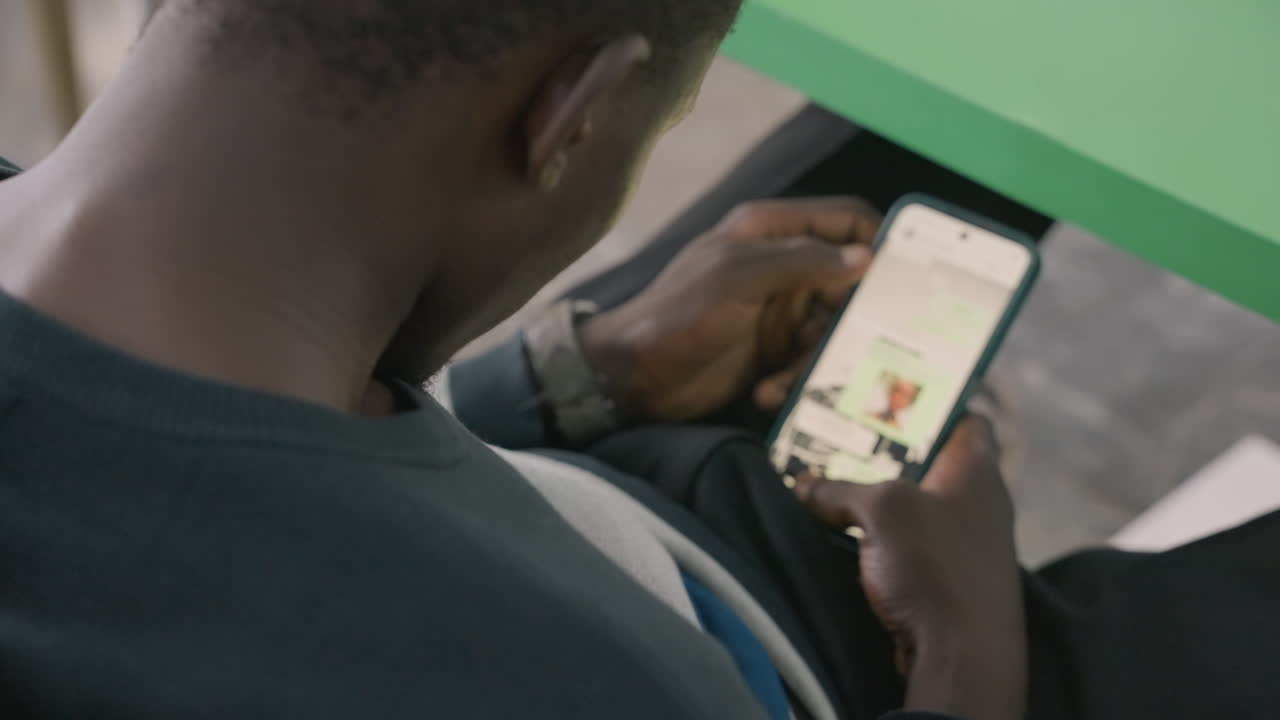 Black man scrolling dating app on phone, sitting at green table, checking messages and profiles, fingers tapping screen, app icons visible, casual sweater, candid focused moment, planning meetup