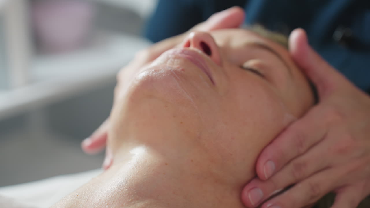 Visitor lies relaxed with eyes closed while beauty expert massages product across cheeks and forehead, enhancing hydrated glow and promoting calm in serene spa treatment environment
