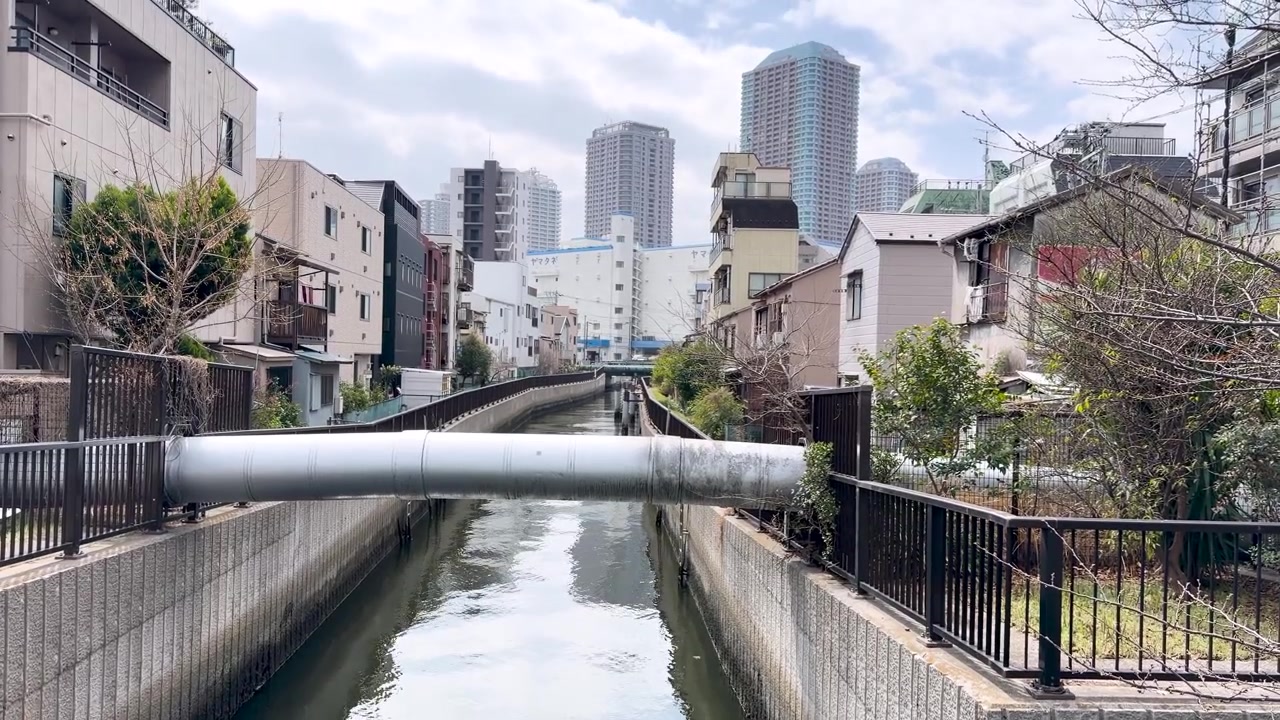 Bir kamera, beton bir kanaldan yukarı doğru eğilerek Tokyo'nun bir yerleşim bölgesinin silüetini gözler önüne seriyor.