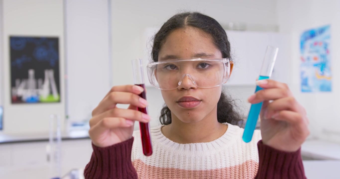 Estudante adolescente usando óculos de proteção, comparando tubos de ensaio vermelhos e azuis no laboratório após a indicação do professor, explicando.