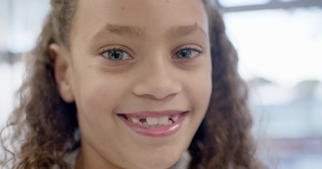 Reacting to camera's eye contact, female child smiling slowly by window, revealing missing tooth
