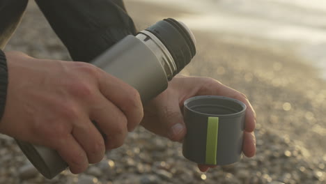 man opens thermos, pours coffee at beach with sea waves golden bay blurry background