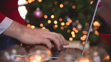 vista de cerca de las manos del hombre escribiendo en la computadora portátil sentada cerca del árbol de navidad en la sala de estar con decoración navideña 1