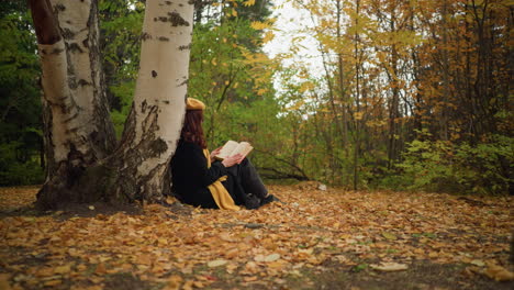 young lady in yellow beret with red hair cascading down her shoulder carefully reading book in hand while seated against a birch tree, surrounded by autumn leaves in peaceful solitude