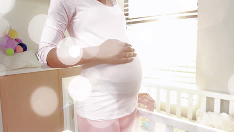 manchas de luz bokeh sobre la sección media de una mujer biracial embarazada frotando el vientre junto a la cuna