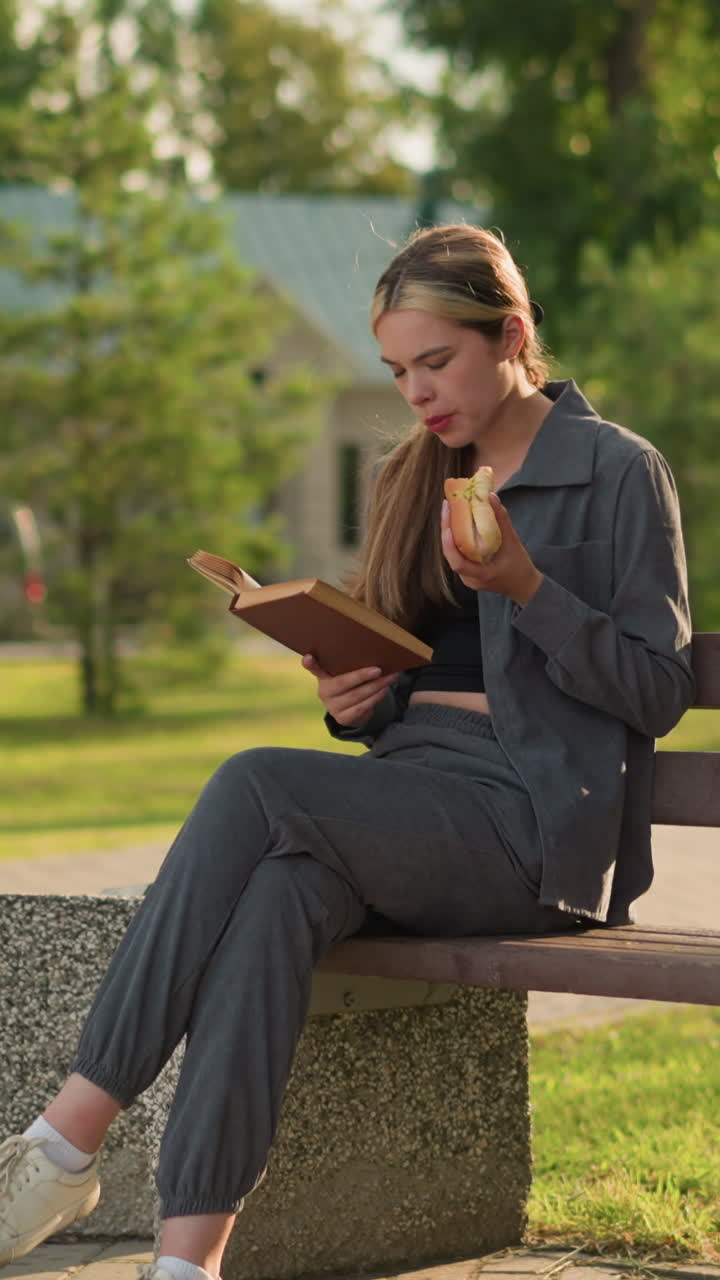 Lady eating a snack while reading book outdoors, seated on park bench with crossed leg, surrounded by greenery, trees, and a building in the background