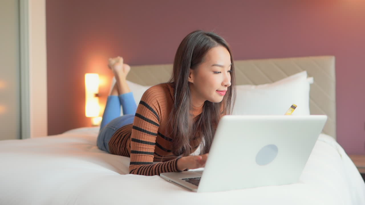 아름다운 아시아 여성이 노트북을 앞에 두고 침대에 누워 음식 배달비를 지불하기 위해 신용카드 번호를 입력합니다.