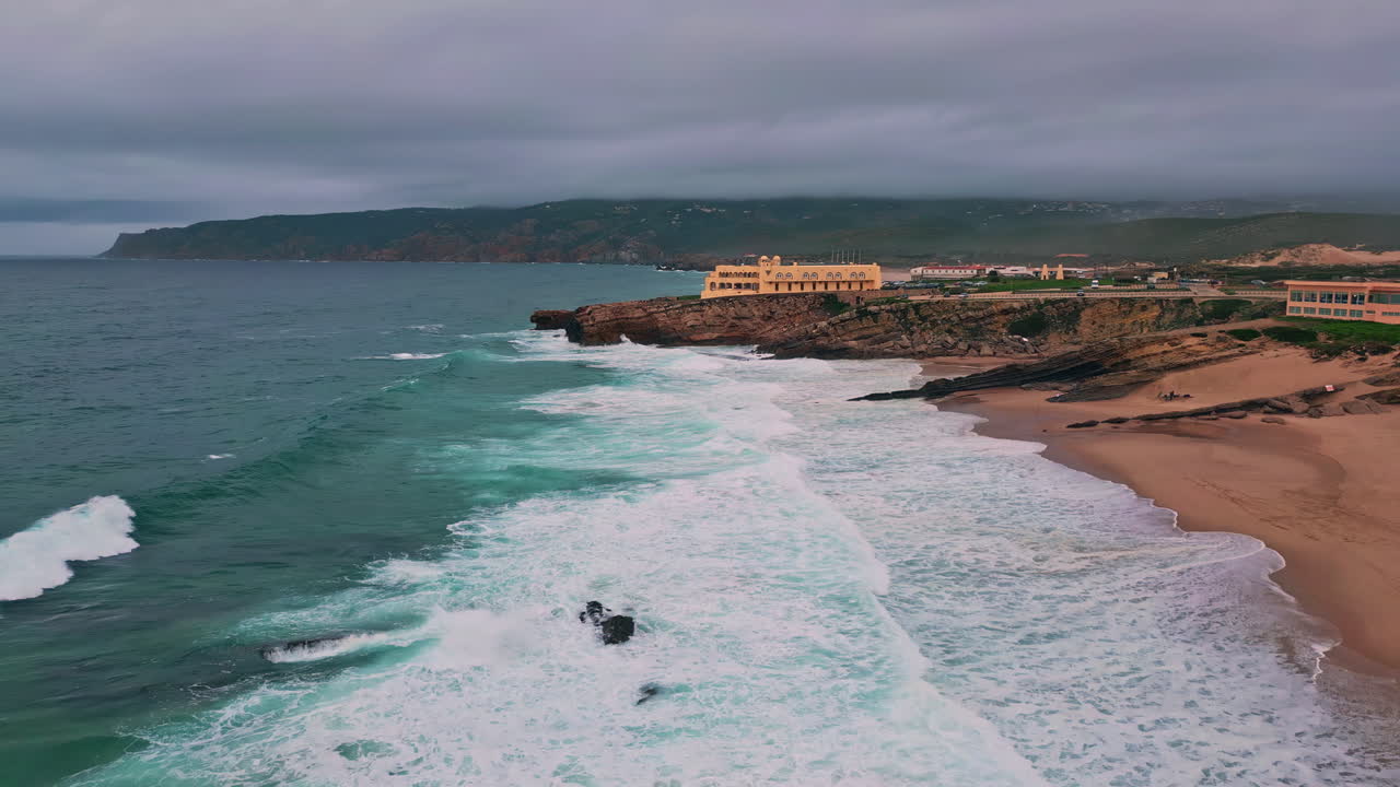 Cliffside hotel overlooking foamy waves at serene beach dusk. Drone view coast
