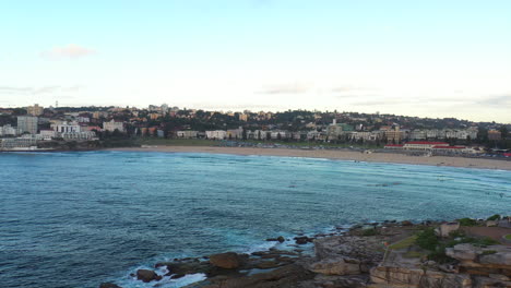 Drone-Volando-A-Través-De-Bondi-Bay-Hacia-La-Playa-Mientras-La-Gente-Rema-En-El-Agua,-Sydney,-Australia