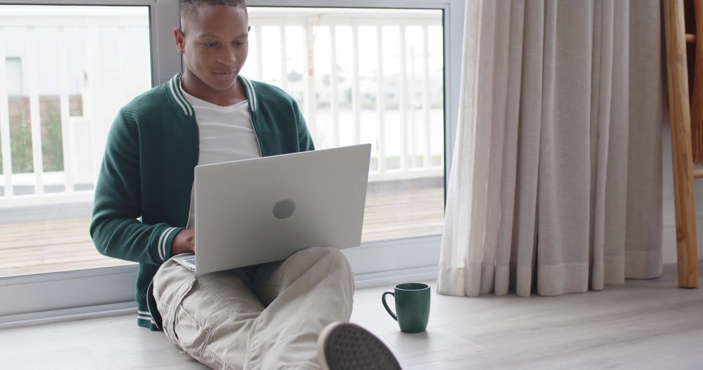 Adult African American man sitting on floor working on laptop, using balcony light, mug nearby