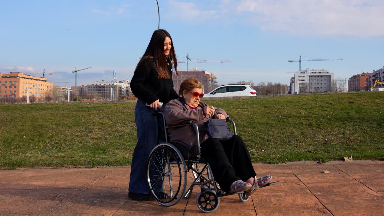 Elderly woman laughing in wheelchair outside with young woman while applying perfume on a sunny day