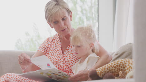 grandmother sitting on sofa with granddaughter at home reading book together