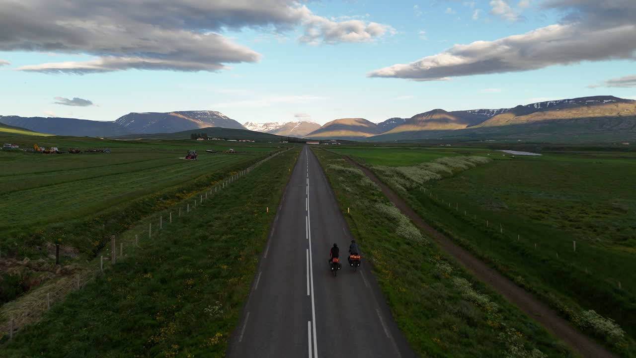 Luftfoto med drone af to cykelturister, der cykler langs vej 76 nær Hofsós i den gyldne time, med dramatiske skyer og varmt solnedgangslys over grønt landbrugsland og Skagafjörður-fjorden på Island