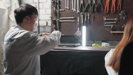 inventor seated with colleague picks up protective glasses, puts them on under fluorescent light, then picks up a tablet, surrounded by tools and equipment in automotive mechanical workshop setting