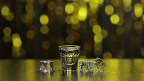 El-Barman-Vierte-Vodka-Congelado-De-La-Botella-En-Un-Vaso-De-Chupito.-Cubitos-De-Hielo-Sobre-Fondo-De-Fiesta-Dorado-Brillante