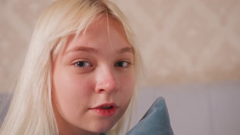 little girl with long blonde hair sits calmly holding soft blue pillow close to face while looking at camera, indoor setting with gentle lighting and neutral background