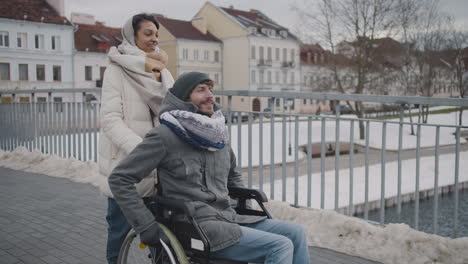 Glückliche-Muslimische-Frau,-Die-Im-Winter-2-Ihren-Behinderten-Freund-Im-Rollstuhl-Auf-Einem-Spaziergang-Durch-Die-Stadt-Mitnimmt-2