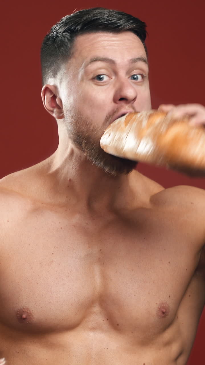 Hungry strong male eating unhealthy food. Half-length portrait of muscular man without shirt eats bread and sausage quickly on the studio background. Vertical video