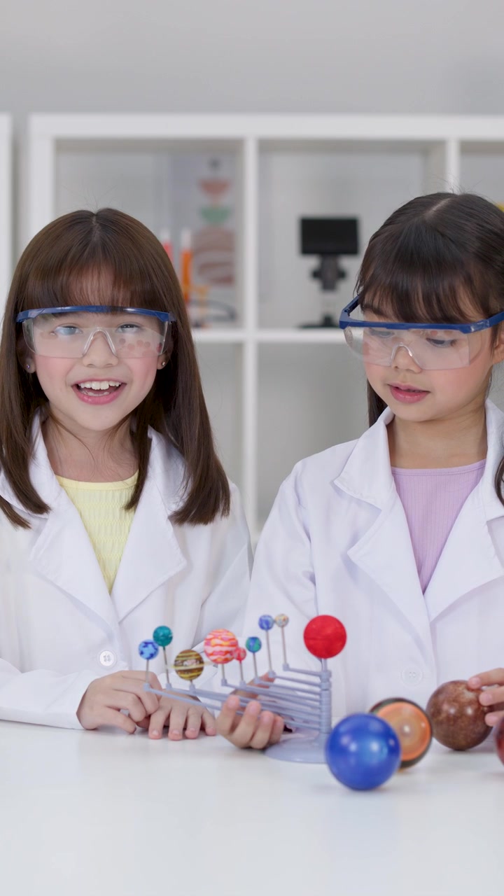 Two young girls in safety goggles examine a planetary model during a school science project.