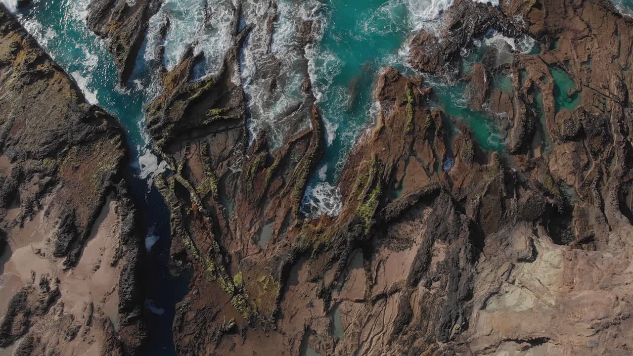 scendendo lentamente verso terra si rivela la superficie ruvida delle rocce in riva al mare in contrasto con le acque turchesi limpide e turbolente