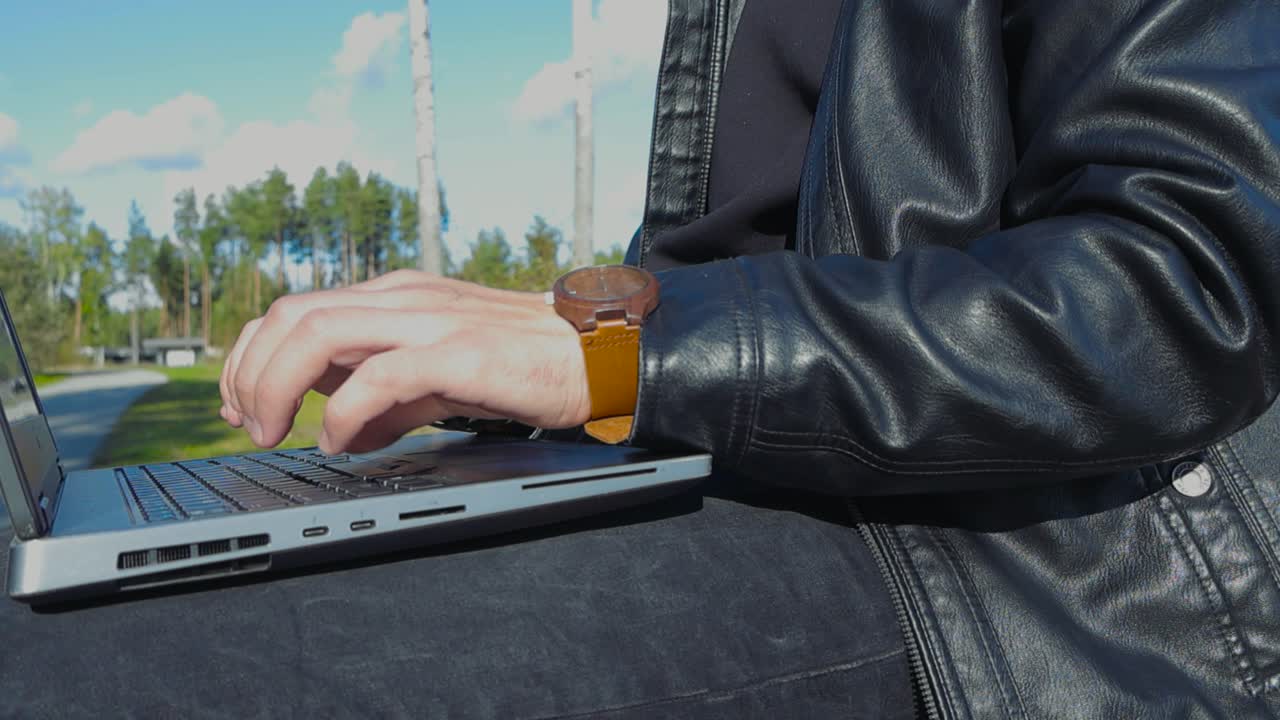 Close up slow motion footage of a person working, studying and typing on a gray laptop and closing it after finishing outdoors while the sun is shining it the park he is working at. Hands in focus.