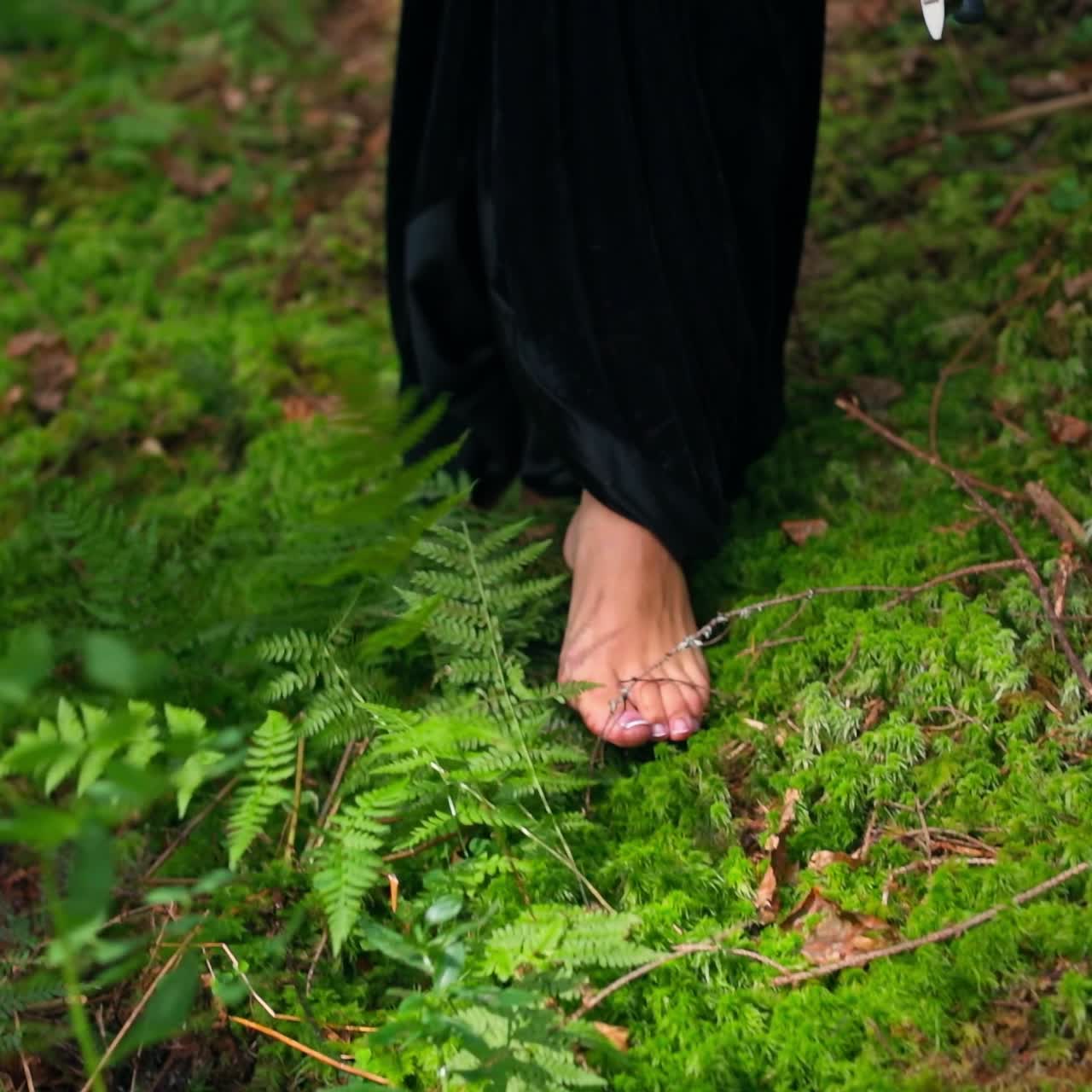 Bare female's feet. Legs of a woman in long black dress walking on green soft grass. Close-up. Slow motion.