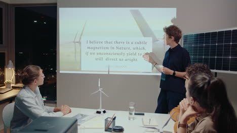 joven empresaria señalando a la pantalla de proyección, dando una presentación sobre recursos de energía sostenible, celebrando una reunión de equipo en la oficina por la noche