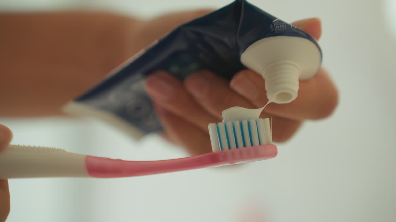 Closeup toothpaste squeezed onto toothbrush hotel bathroom, guest hand applies mint paste onto pink bristles, soft natural light, shallow depth of field, minimal aesthetic, intimate hygiene routine
