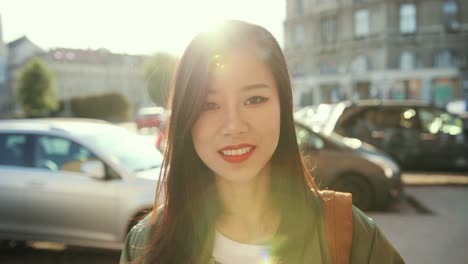 Portrait-Shot-Of-The-Charming-Youung-Stylish-Woman-Smiling-Happily-To-The-Camera-And-Fixing-Her-Hair-With-A-Hand-At-The-Noisy-Street-With-Traffic