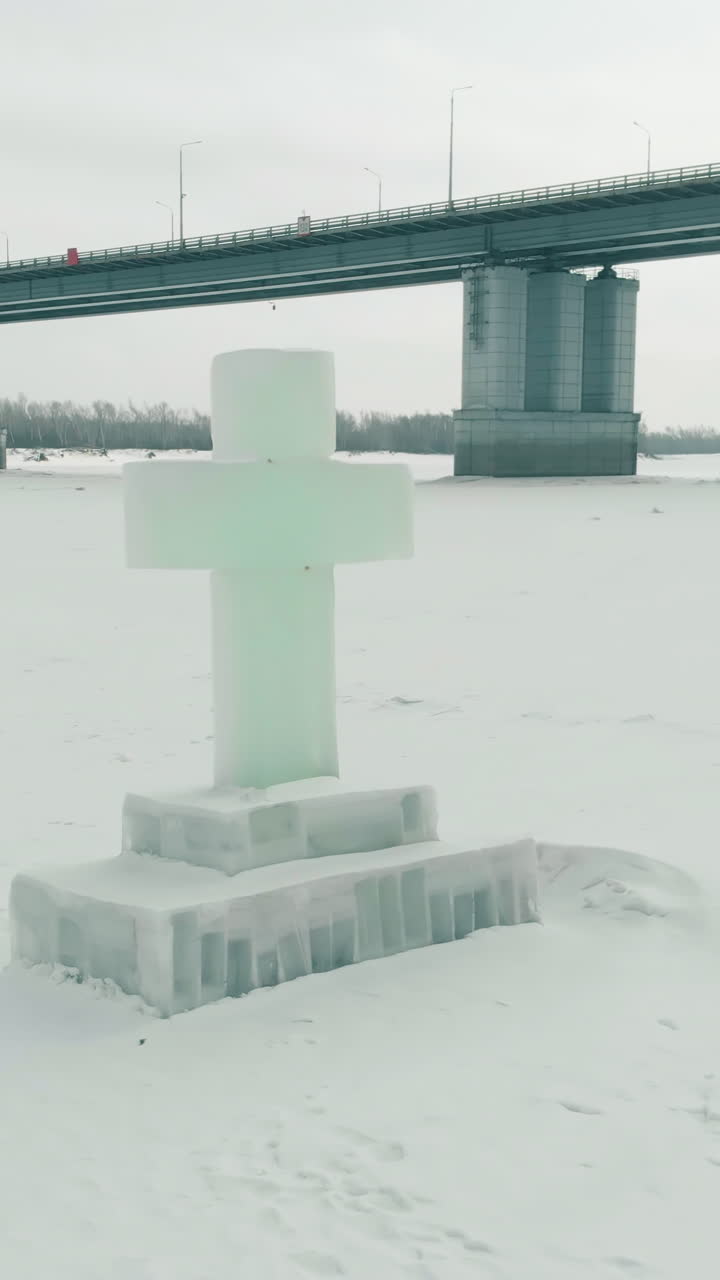 carved large ice cross for Epiphany holiday on white frozen river against huge bridge and people silhouettes upper view