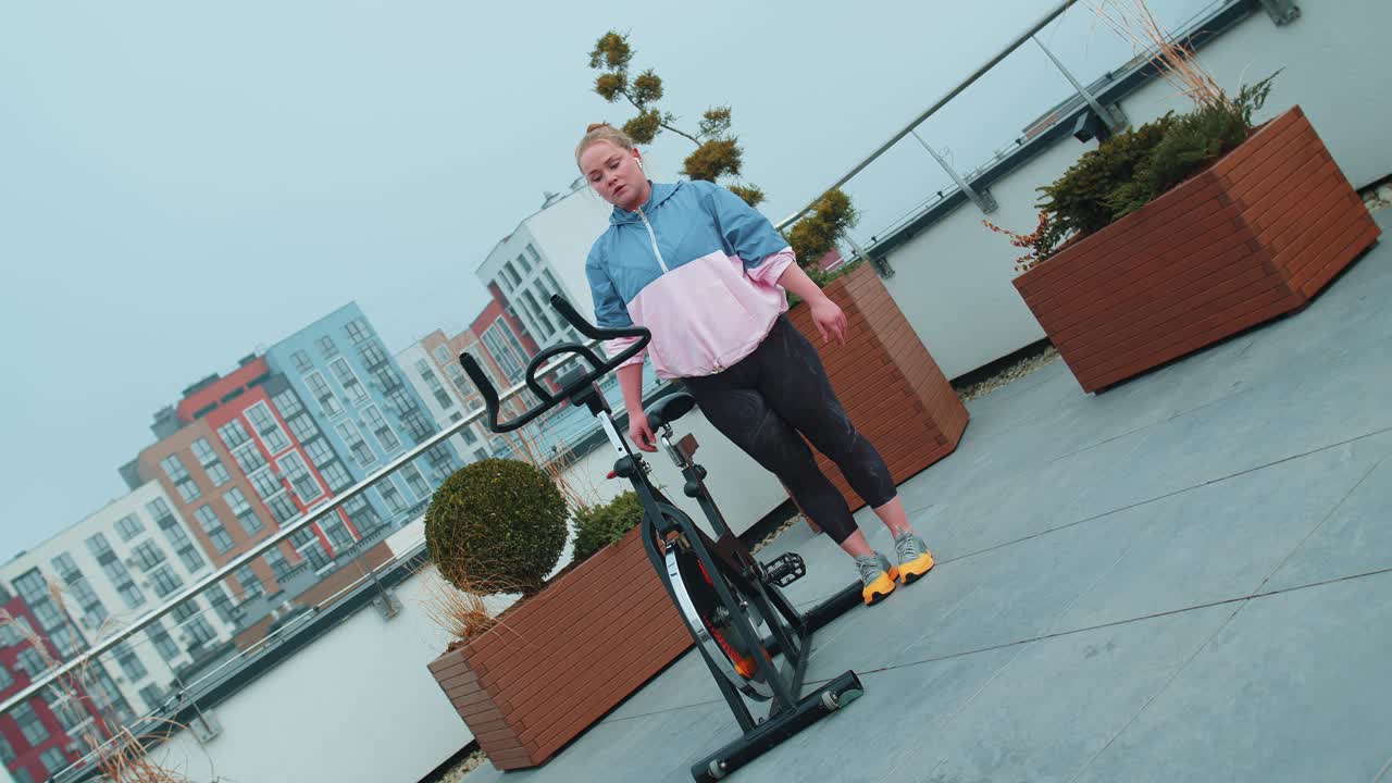 Athletic girl performing aerobic training twine exercises on cycling stationary bike on house roof