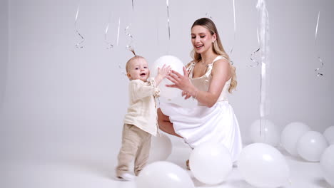 beautiful caucasian woman sits squatted calling her baby to come up. adorable infant making first steps to approach his mommy. lovely kid grabs a balloon to play. studio footage.