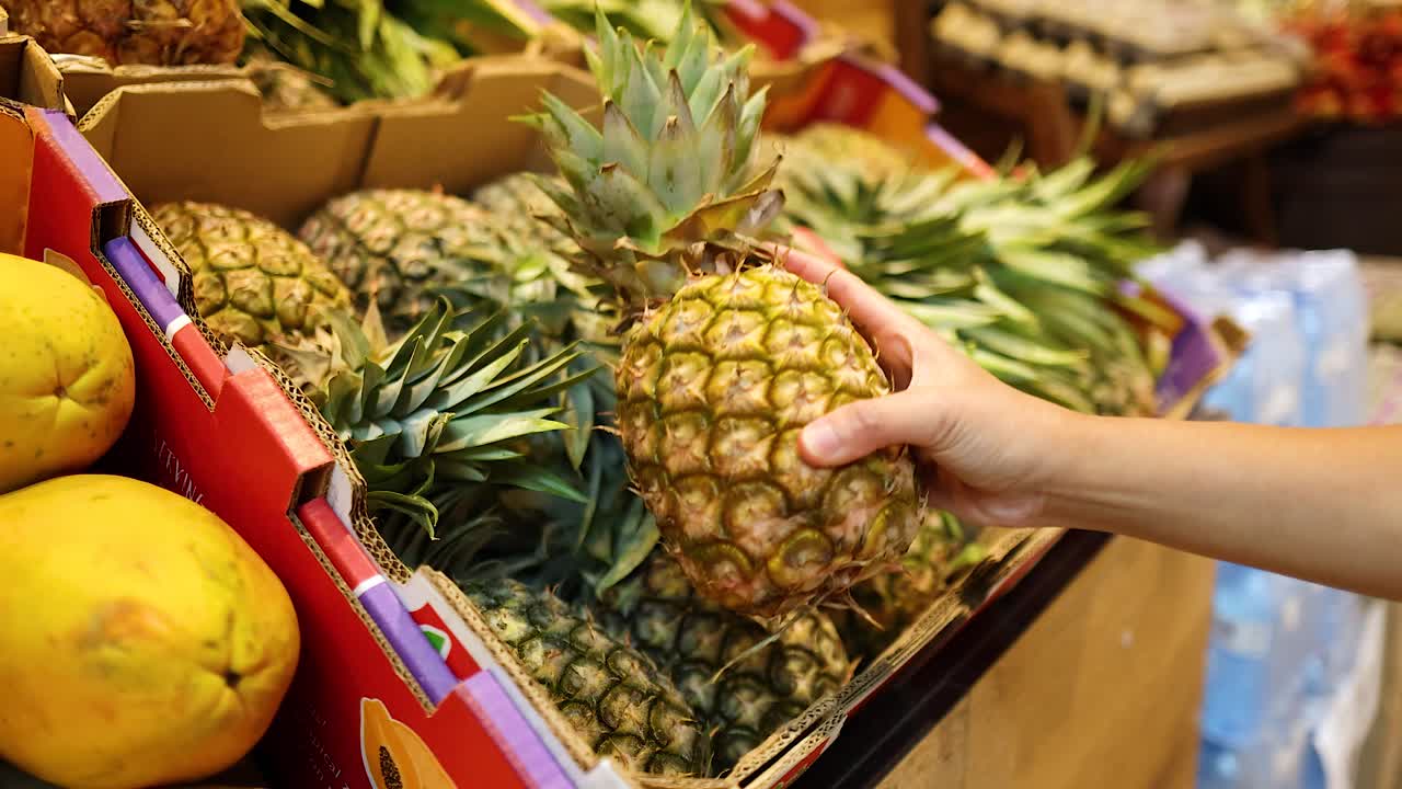 A hand reaches for a pineapple in a vibrant supermarket setting, showcasing fresh produce under bright lighting