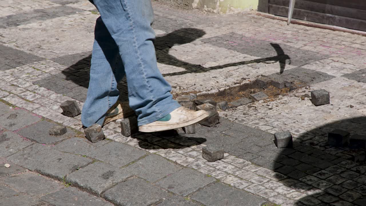 Construction worker uses pickaxe to lift cobblestones on sunlit Prague sidewalk, static camera view