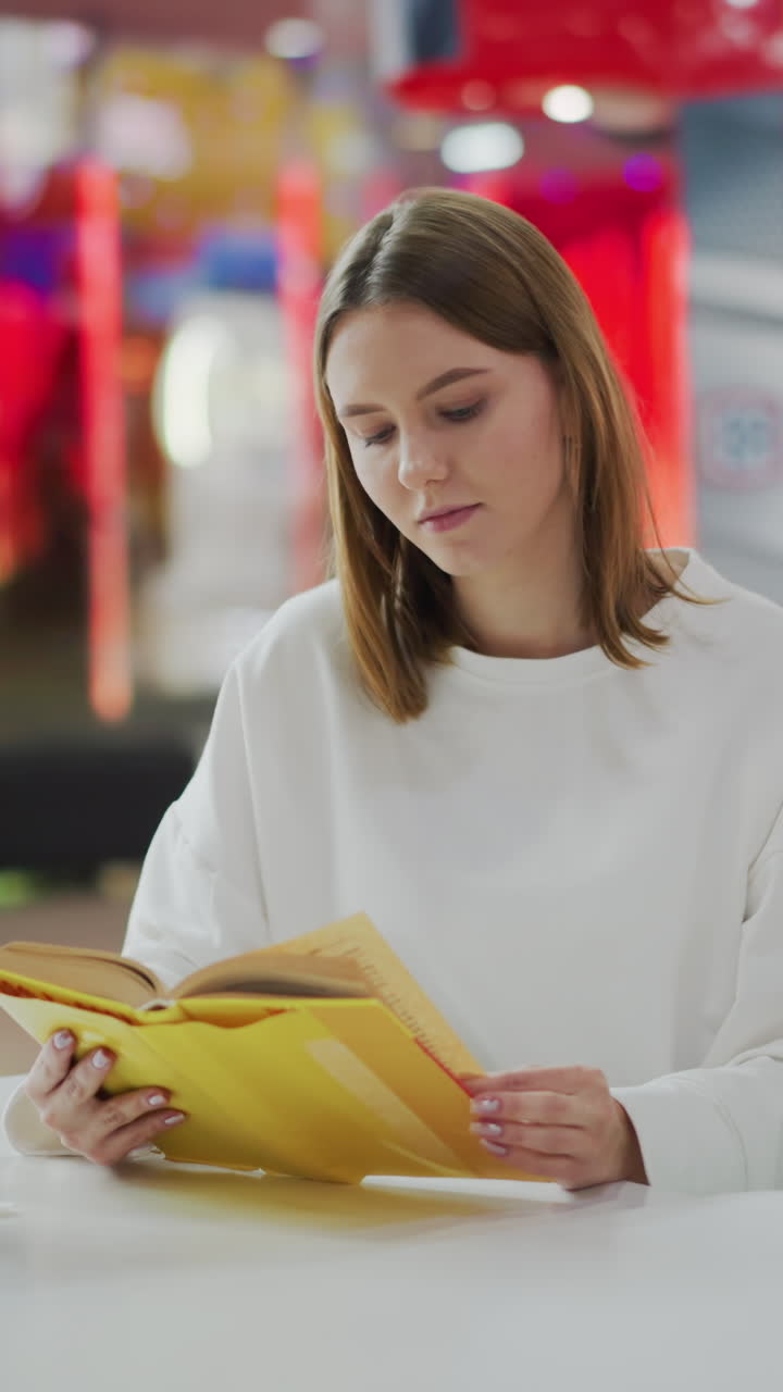 mujer sentada en un centro comercial leyendo un libro amarillo, con taza de café y bolsas de compras de colores en la mesa, rodeada de iluminación suave, decoración vibrante y fondo borroso de juguetes y compradores